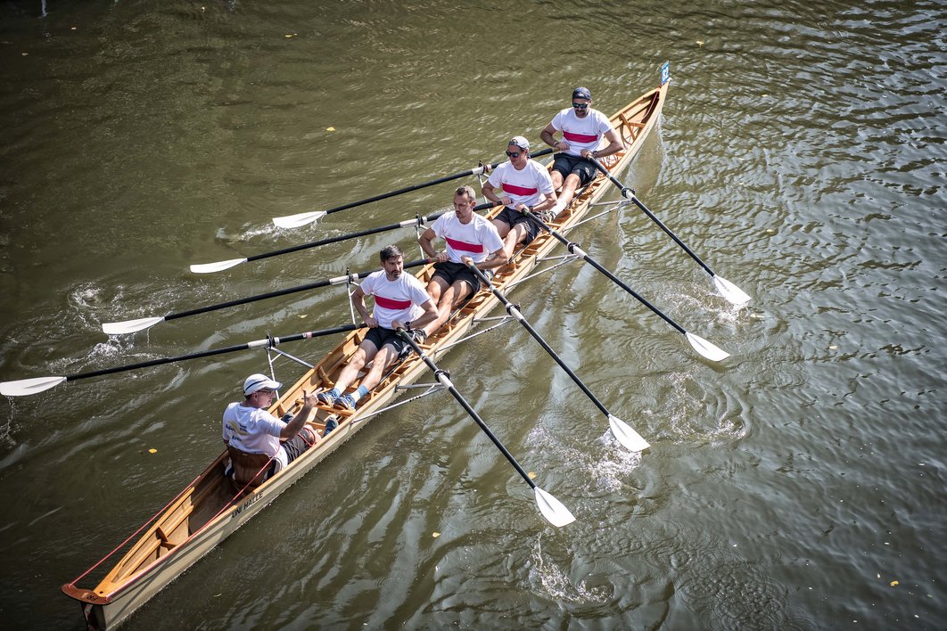 Vier Personen und ein Steuermann rudern in einem Ruderboot auf der Saale in Halle. Das Foto wurde von oben aufgenommen, das Boot rudert diagonal durchs Bild.