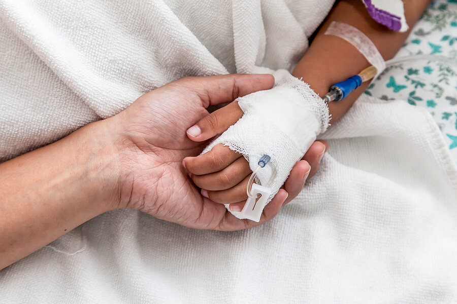 Detailaufnahme: Die Hand eines Kindes liegt in der Hand einer erwachsenen Person. In der Hand des Kindes befindet sich ein Zugang mit einer Flexüle. Detailaufnahme: Die Hand eines Kindes liegt in der Hand einer erwachsenen Person. In der Hand des Kindes befindet sich ein Zugang mit einer Flexüle.