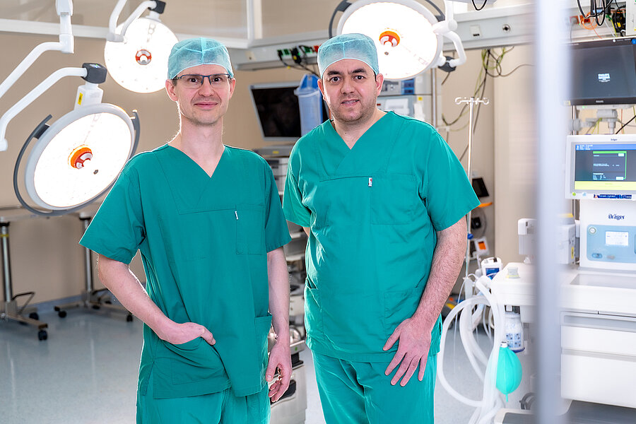 Zwei Personen in OP-Kleidung stehen in einem Operationssaal. Im Hintergrund und rechts im Bild befinden sich Lampen und andere OP-typische Geräte. Zwei Personen in OP-Kleidung stehen in einem Operationssaal. Im Hintergrund und rechts im Bild befinden sich Lampen und andere OP-typische Geräte.
