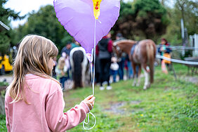 Ein Kind mit blonden Haaren und rosa Pullover steht im Vordergrund und hält einen lilafarbenen Luftballon in Herzform. Im Hintergrund sind mehrere Menschen und zwei Ponys zu sehen, die auf einer Wiese stehen.