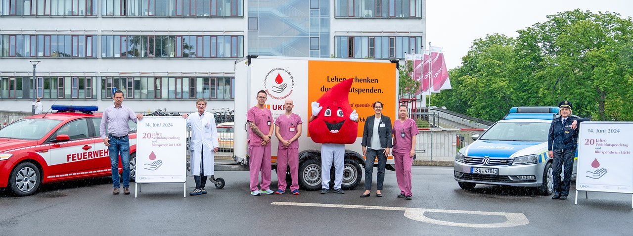 Gruppenbild auf dem 2. Hubschrauberlandeplatz vor dem UKH: Auf dem Platz stehen je ein Fahr-zeug der Feuerwehr und der Polizei sowie ein Anhänger mit Werbung für die Blutspende des UKH. Vor den Autos und Anhängern stehen die Vertreter:innen der Feuerwehr und der Polizei sowie Mitarbeitende der Blutspende und der Notaufnahme des UKH. In der Mitte steht das Maskottchen der Blutspende, ein großer Bluttropfen.