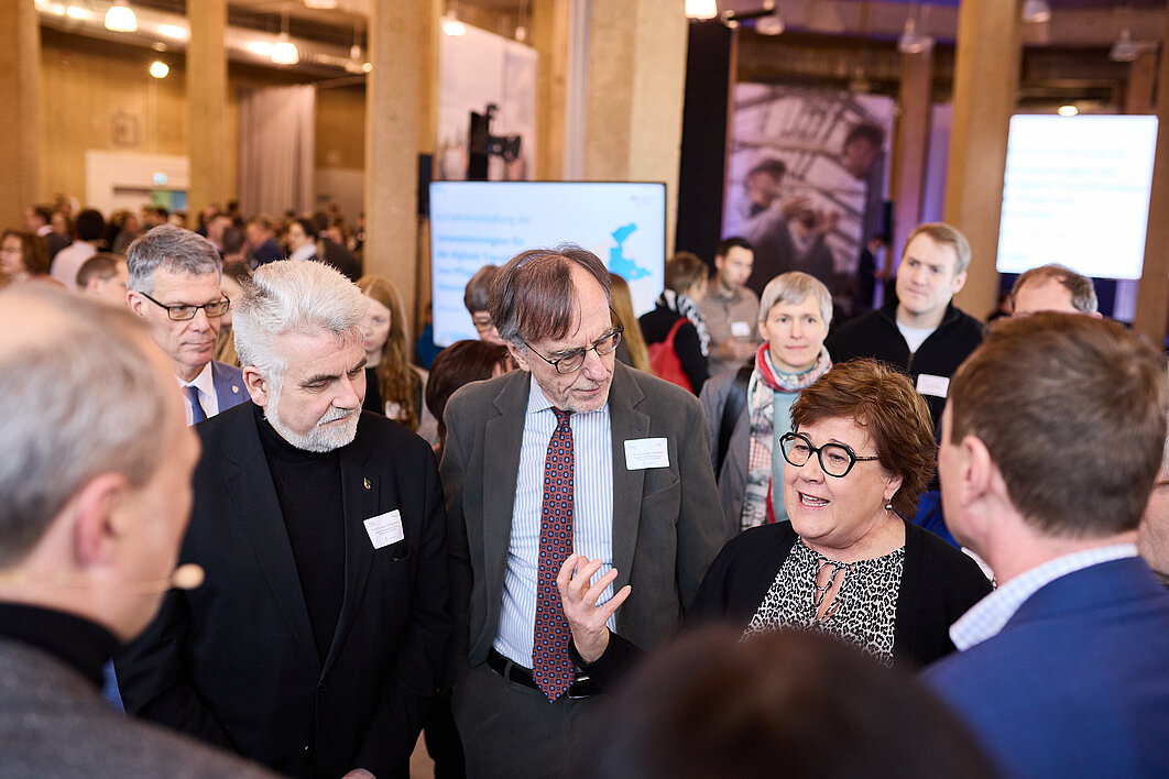 Es sind im Kreis stehende Menschen zu sehen. Direkt im Vordergrund sind nur die Hinterköpfe zu sehen. Der Fokus liegt auf einer Gruppe Menschen, die von vorn zu sehen sind. Eine Frau rechts erläutert gerade mit einer Handgeste etwas. Die beiden links daneben stehenden älteren Männer schauen sie zuhörend an.