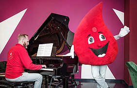 Eine Person sitzt im Foyer des Neues Theaters in Halle (Saale) an einem Flügel. Rechts neben dem Flügel steht eine Person im Maskottchen-Kostüm der Blutspende der Universitätsmedizin Halle und winkt. Das Maskottchen-Kostüm ist ein großer Blutstropfen.