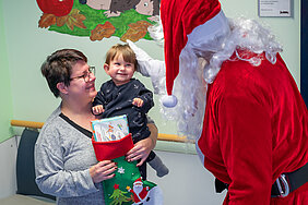 Ein kleines Kind sitzt auf dem Arm seiner Mutter und lacht einen Weihnachtsmann an, der ihm den Kopf streichelt. Der Weihnachtsmann ist von schräg hinten zu sehen. Die Mutter hält einen Weihnachtssocken mit Geschenken in den Händen.