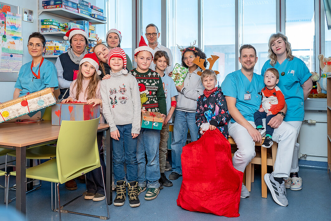 Gruppenbild von einer Gruppe Kinder und sechs Erwachsenen. Die Gruppe befindet sich im Spielzimmer einer Kinderstation eines Krankenhauses. Einige Personen tragen Weihnachtsmützen und halten Geschenke in den Händen. Ein Kind hält einen großen roten Sack mit Geschenken fest.