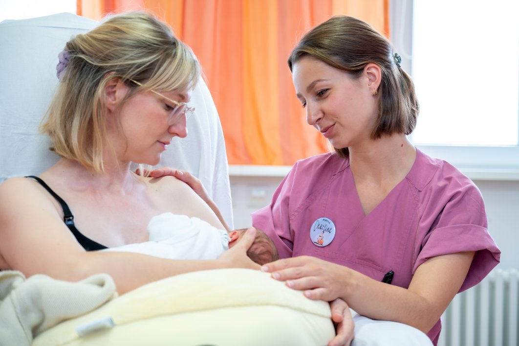 Eine Hebamme sitzt neben einer Mutter, die stillt. Beide schauen auf das Baby, das auf einem Stillkissen auf dem Schoß der Mutter liegt.