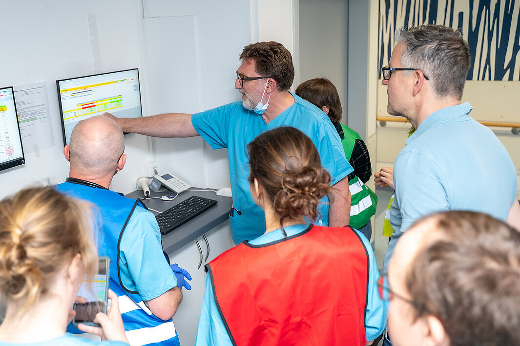 Mehrere Personen stehen um einen Monitor herum. Einige tragen medizinische Arbeitskleidung. Eine Person zeigt etwas auf dem Monitor.