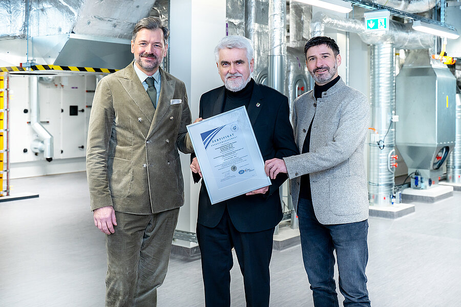 Drei Männer in Anzügen halten gemeinsam ein Zertifikat. Im Hintergrund ist ein Technikraum zu sehen. Drei Männer in Anzügen halten gemeinsam ein Zertifikat. Im Hintergrund ist ein Technikraum zu sehen.