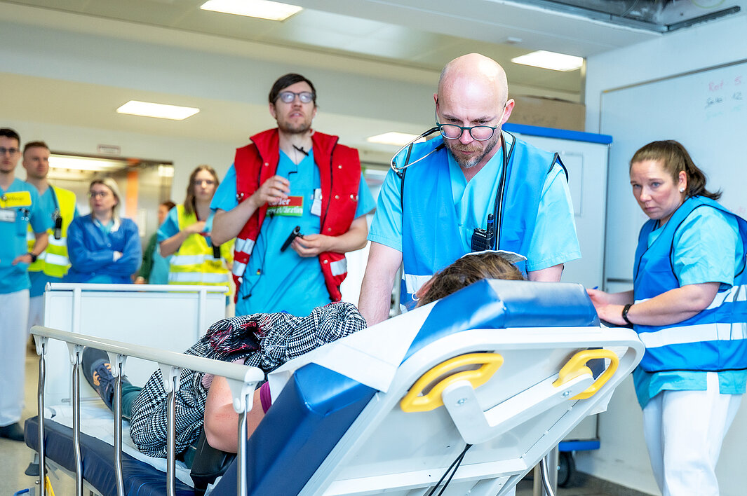 Eine Person in medizinischer Arbeitskleidung versorgt eine Person, die auf einer Trage liegt. Im Hintergrund befinden sich weitere Personen in medizinischer Arbeitskleidung.