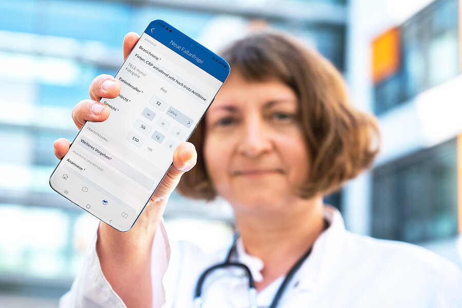 Eine Person in medizinischer Arbeitskleidung hält ein Smartphone in die Kamera. Dieses zeigt die geöffnete Telekasper-App. Eine Person in medizinischer Arbeitskleidung hält ein Smartphone in die Kamera. Dieses zeigt die geöffnete Telekasper-App.
